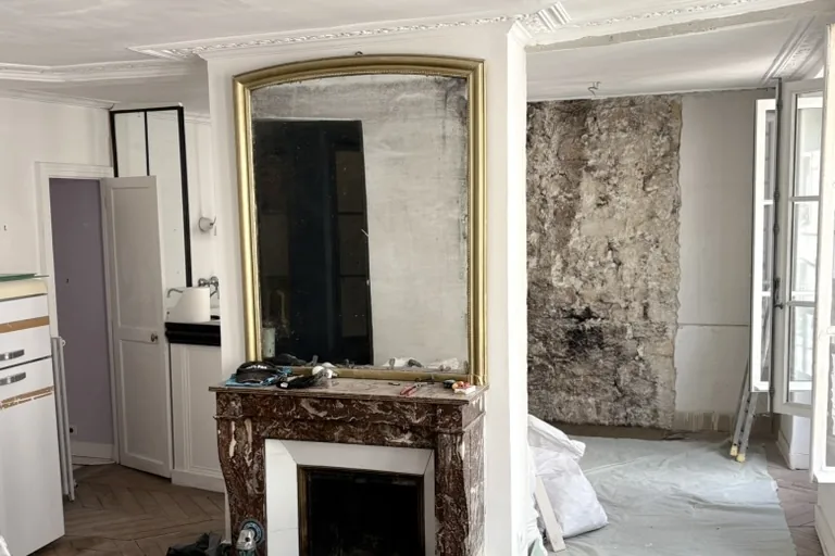 Appartement haussmannien parisien en cours de rénovation : parquet à point de Hongrie, cheminée en marbre et trumeau doré, mur mis à nu, bâches de protection ; espace cuisine à repenser dans le bâti ancien.