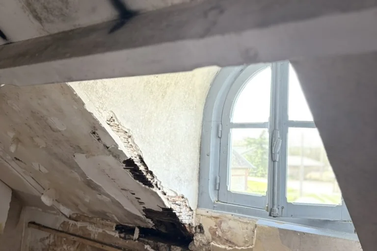 Sous-comble d'un manoir normand du XVIIe siècle : fenêtre cintrée, plafond en lattis et plâtre ouvert par une infiltration d'eau, charpente et mur de brique mis à nu.
