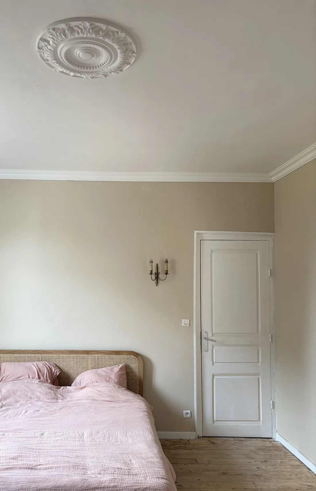 Suite parentale terminée dans un manoir : murs greige, tête de lit en cannage, linge de lit rose poudré, applique en laiton et rosace de plâtre restaurée au plafond.
